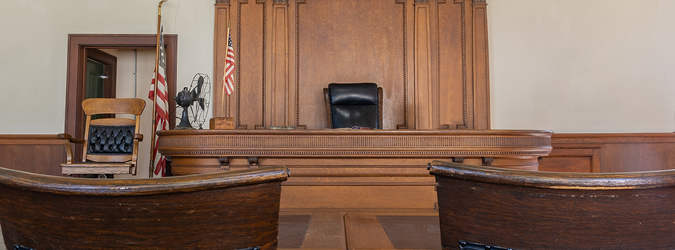 Empty wooden judges bench in a courtroom with two chairs, two American flags, and a large wooden backdrop behind the bench.