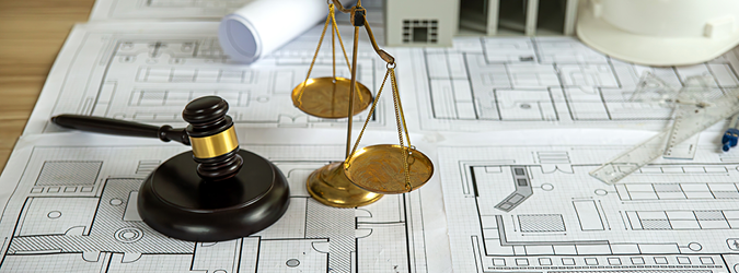 A judges gavel and brass scales of justice rest on architectural blueprints, with drafting tools and building models in the background.