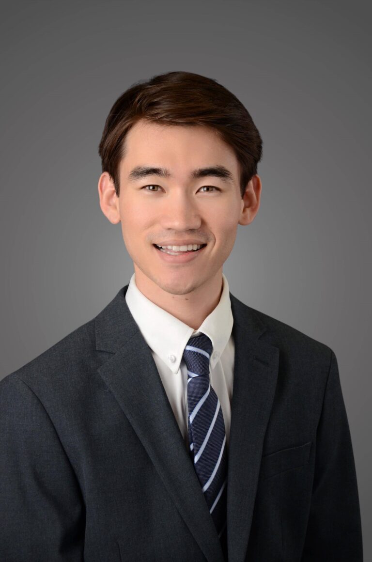 A young man with short brown hair, wearing a dark suit, white shirt, and striped tie, smiles in front of a plain gray background.