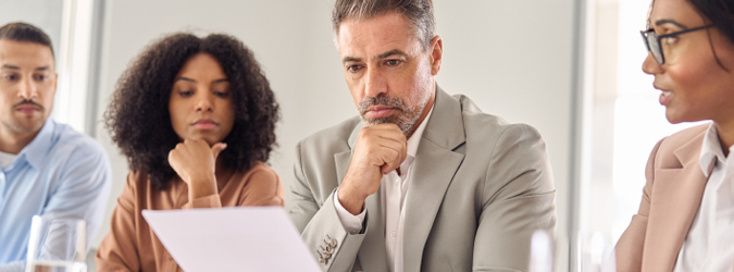 Four people sit at a table, focused on a document one man is holding. They appear to be in a business meeting or discussion, with thoughtful and attentive expressions.