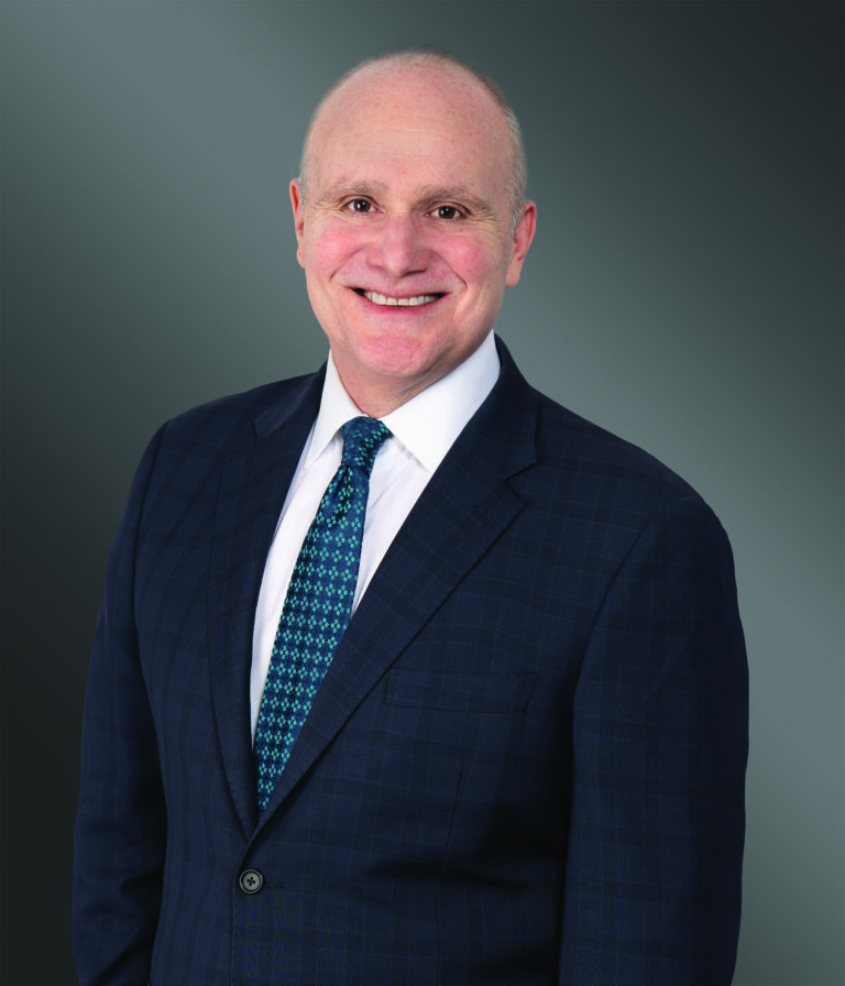 A smiling man in a blue checkered suit, white shirt, and patterned tie stands against a gray gradient background.
