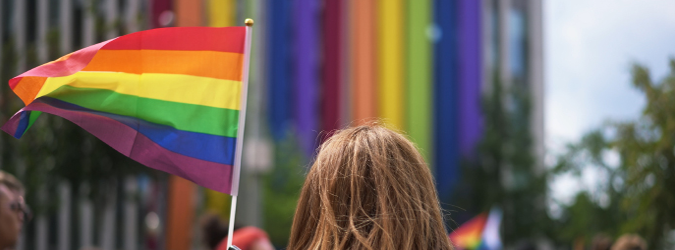 A person with brown hair holds a rainbow pride flag at an outdoor event, with a building decorated in vertical rainbow banners visible in the background.