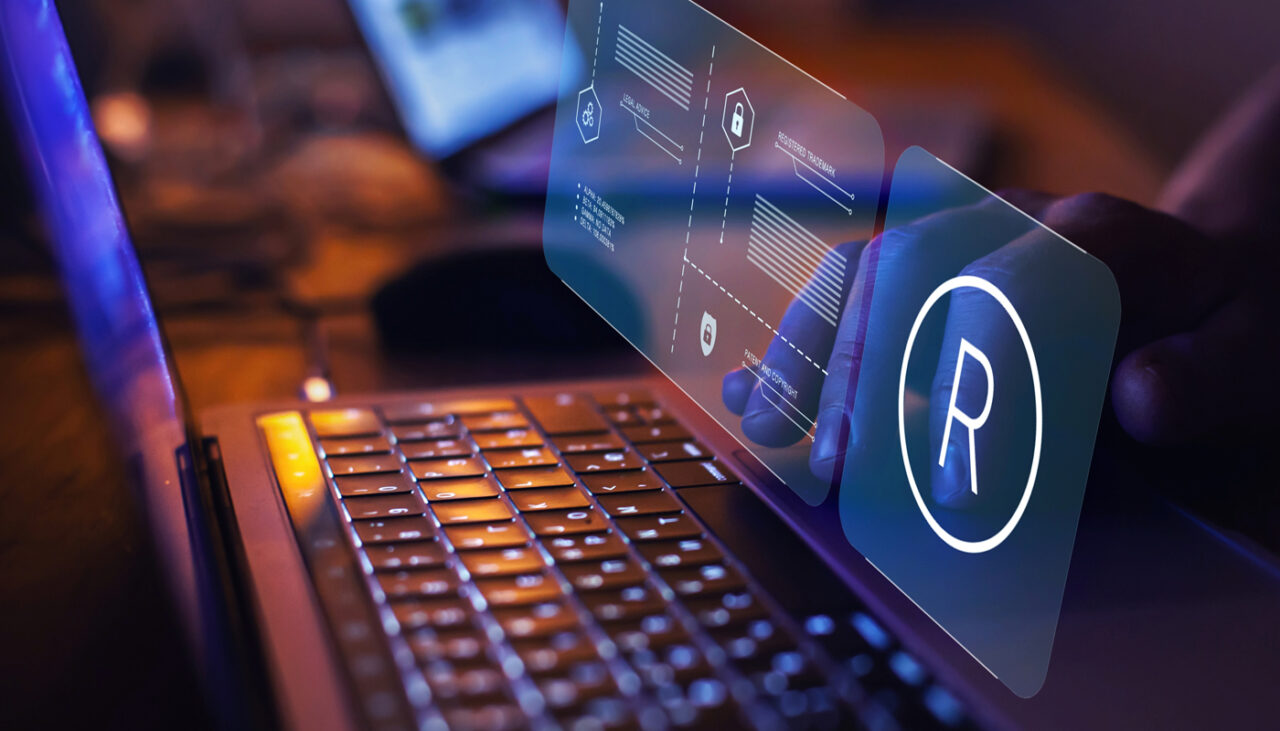 A person types on a laptop with a holographic trademark symbol and digital interface floating above the keyboard, suggesting technology and intellectual property concepts.