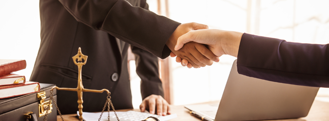 Two people in business attire shake hands across a desk with a laptop, legal books, and a balance scale, suggesting a business or legal agreement.
