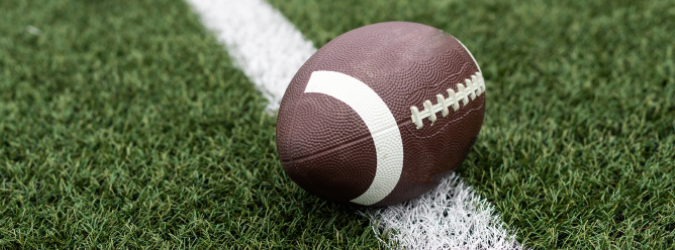 A brown American football with white laces is lying on a green turf field, resting on top of a white sideline.