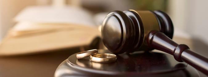 A judges gavel rests beside two gold wedding rings on a wooden block, with an open book blurred in the background.