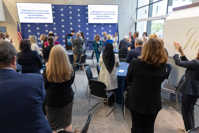 People stand and applaud at an indoor awards ceremony. A blue backdrop displays text, and attendees face the stage where speakers are present. Tables and chairs fill the room, and large windows let in natural light.