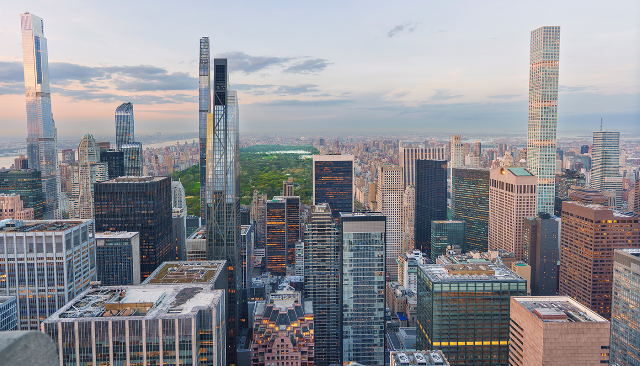 A cityscape of New York Citys Midtown Manhattan with tall skyscrapers in the foreground and Central Park in the background under a partly cloudy sky at sunset.
