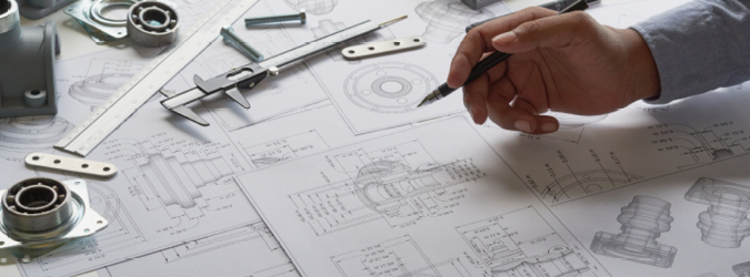 A hand holding a pencil works on detailed mechanical engineering blueprints, surrounded by calipers, metal parts, and a ruler on a cluttered desk.