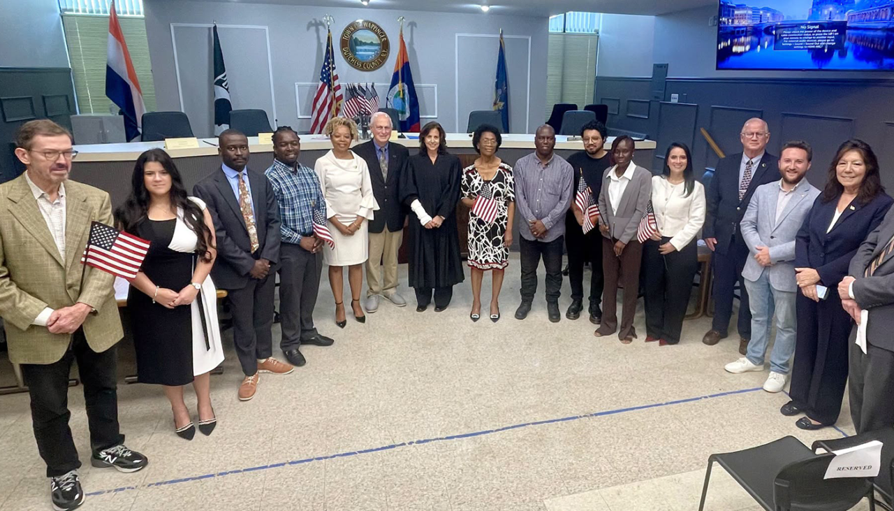 A diverse group of people, some holding small American flags, stand in a government chamber with officials and a judge, suggesting a citizenship or swearing-in ceremony. American flags and official symbols are visible in the background.