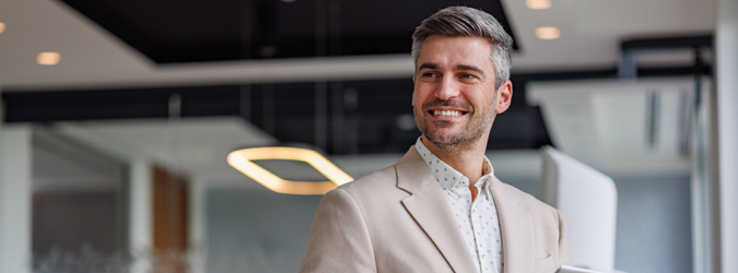 A smiling man with short gray hair, wearing a light beige blazer and white shirt, stands in a modern, well-lit office environment.