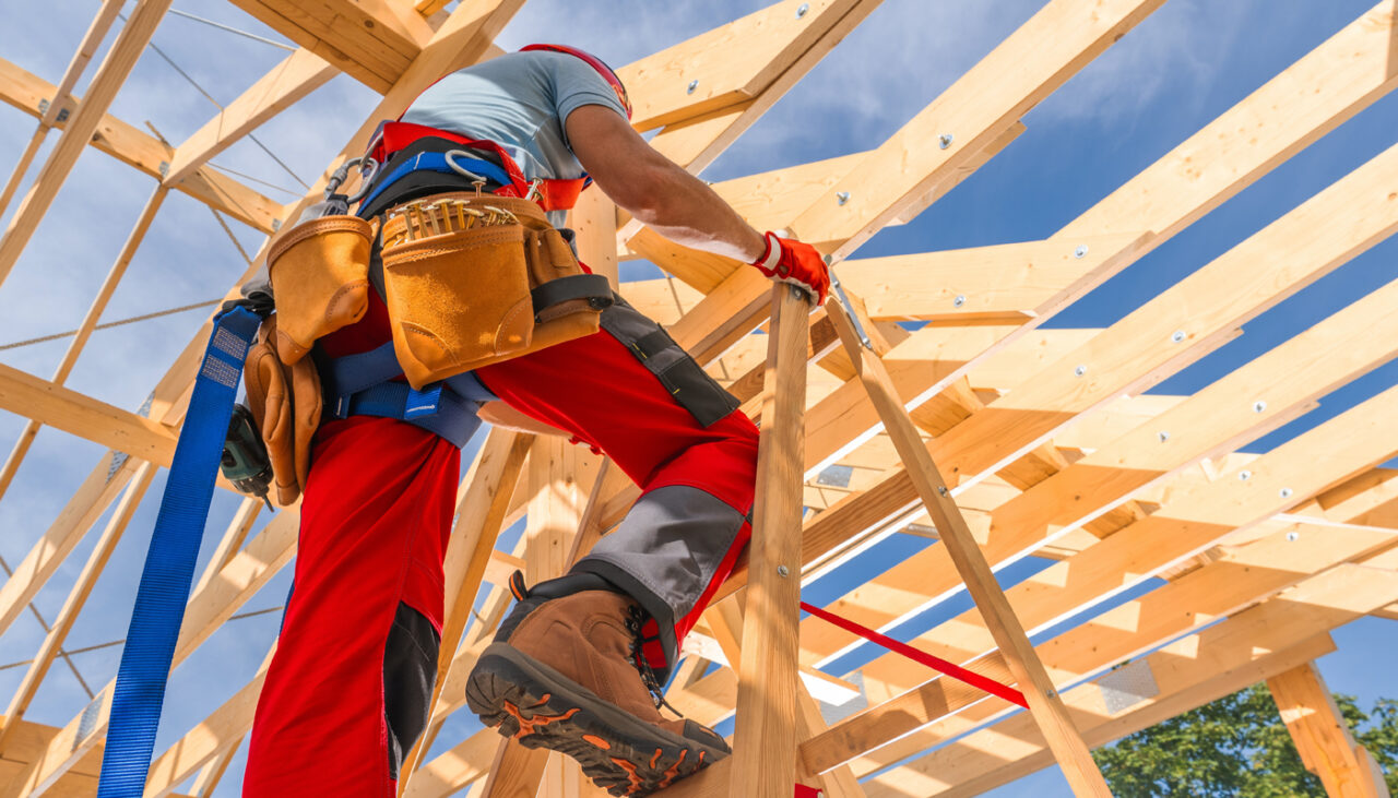 Construction worker in safety gear climbs a wooden frame of a house under construction, with blue sky in the background.