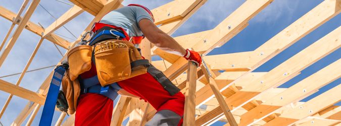 A construction worker wearing a tool belt and safety gear works on a wooden frame of a building under a clear blue sky.