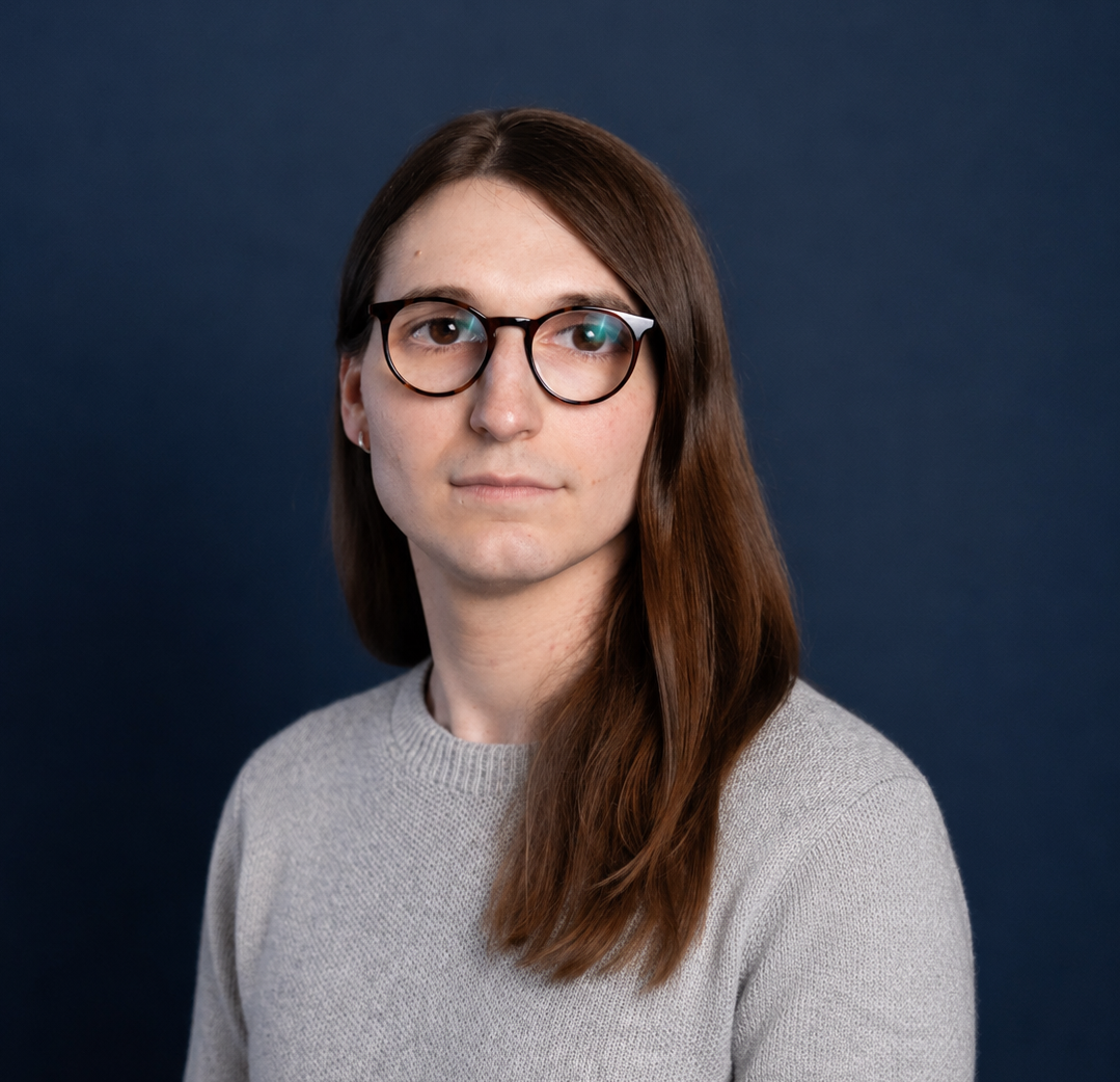 A person with long brown hair, glasses, and a gray sweater looks slightly to the side in front of a plain dark blue background.