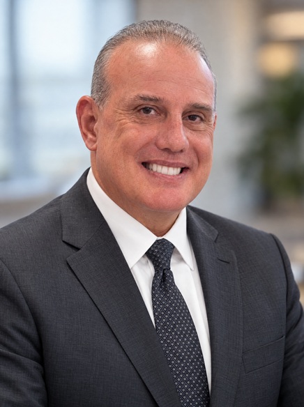 Smiling man in a dark suit, white shirt, and patterned tie, with short gray hair, posing in a bright, blurred office setting.