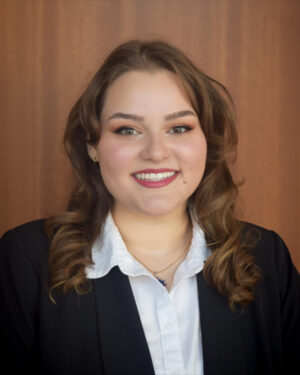 A woman with wavy brown hair, wearing a black blazer and white collared shirt, smiles at the camera in front of a wooden background.