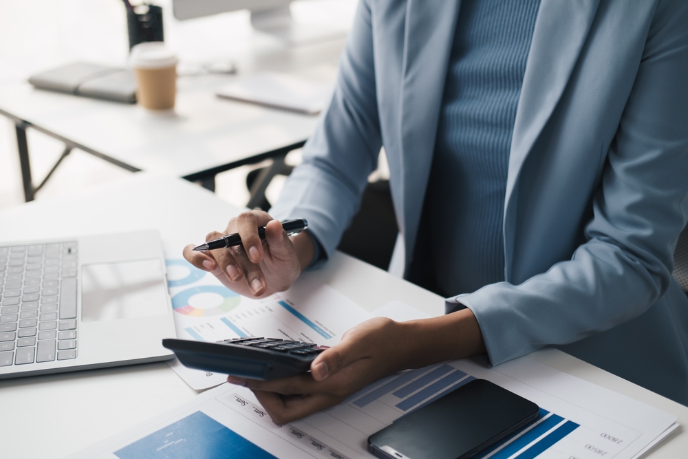 A person in a blue suit uses a calculator and holds a pen at a desk with charts, a laptop, and a smartphone, working on financial documents.