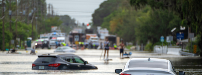 Two people paddleboard down a flooded street past partially submerged cars.