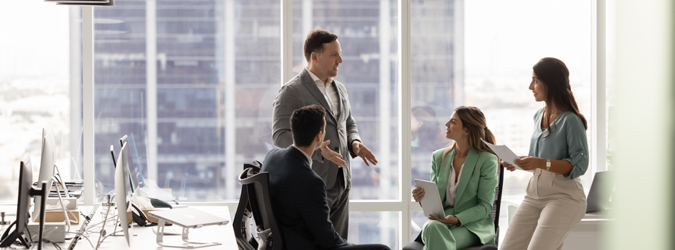 Four professionals have a discussion in a bright office with large windows and city views. Two people are seated, while two stand, all engaged in conversation with documents in hand.