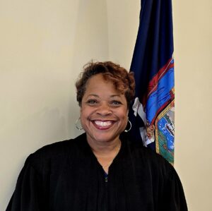 A smiling woman wearing a black judicial robe standing in front of a New York State flag.