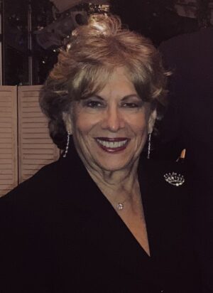 A smiling woman with shoulder-length blonde hair wearing a black blazer, a necklace with a small pendant, and dangle earrings.