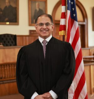 A smiling judge in a black robe and striped tie standing in a courtroom next to an American flag.