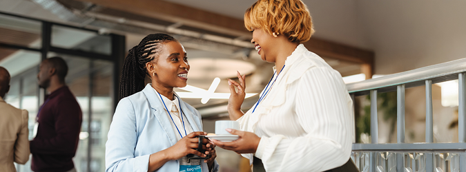 Two professional women with ID badges smile and talk while holding coffee cups in an office hallway.