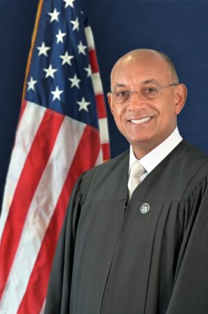 A smiling man wearing glasses and a judges robe standing in front of an American flag.