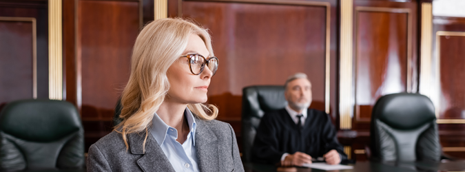 A woman in a grey blazer and glasses sitting in a courtroom with a judge in the background.