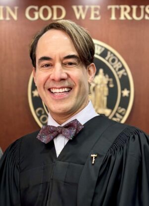 A smiling man wearing a judicial robe and a patterned bowtie, standing in front of a New York state seal.