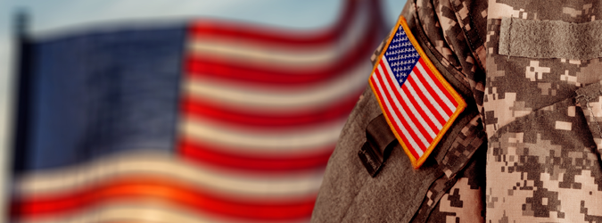 An American flag patch on a camouflage military uniform with a blurred American flag in the background.