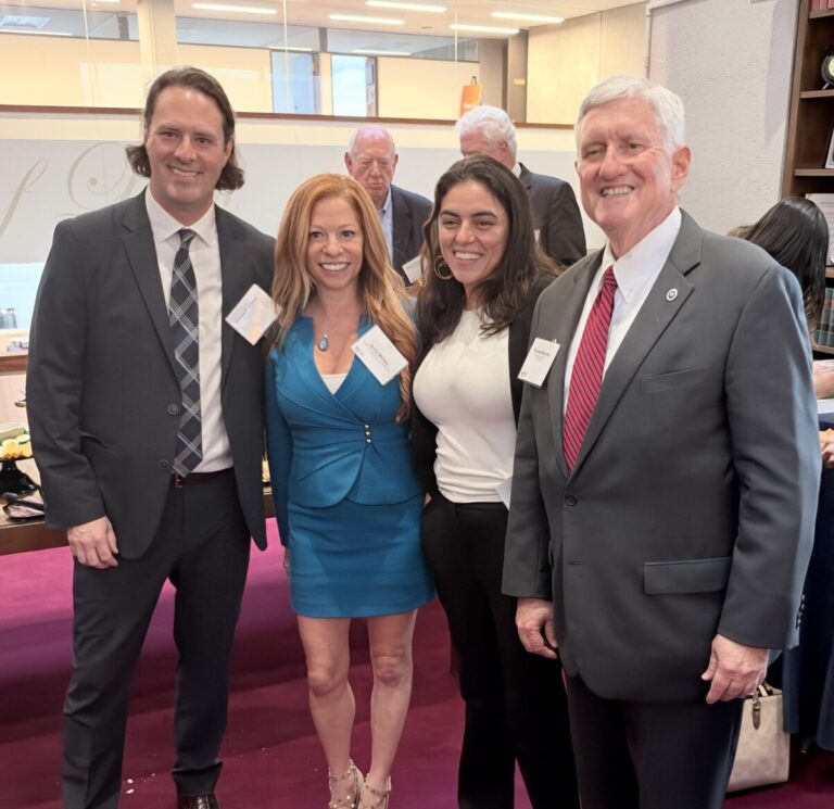 Two men in suits and two women in business attire stand together for a photo in a room with a large wall inscription.