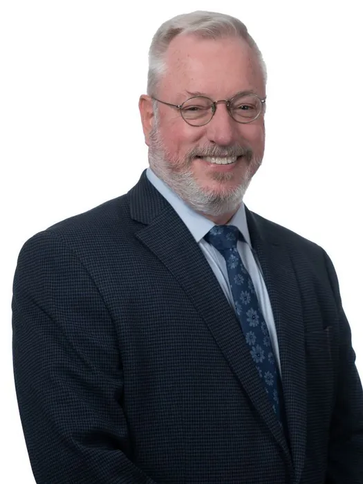 A smiling man with gray hair and glasses wearing a dark suit, light blue dress shirt, and a patterned blue tie.