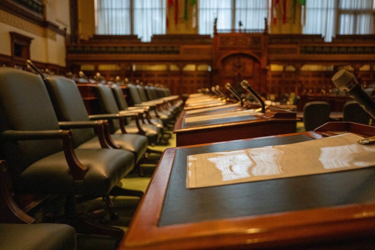 Rows of empty dark leather chairs with microphones line wooden desks in a legislative chamber.