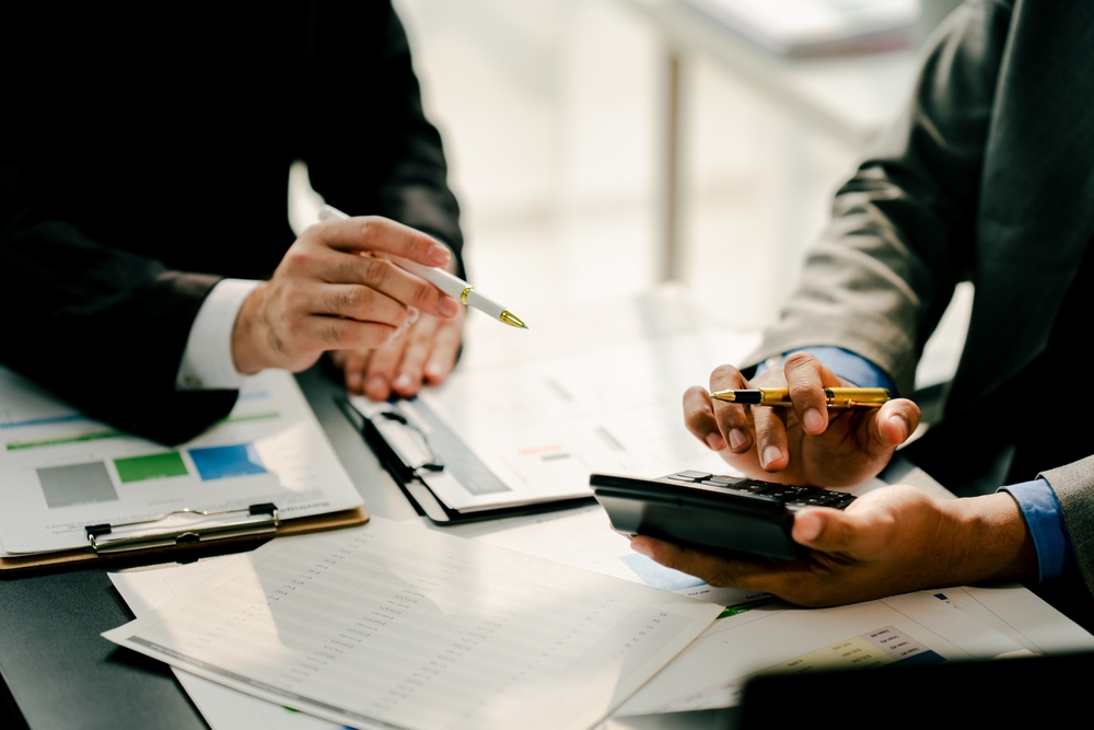 Two professionals in suits review financial documents and use a calculator at a desk.