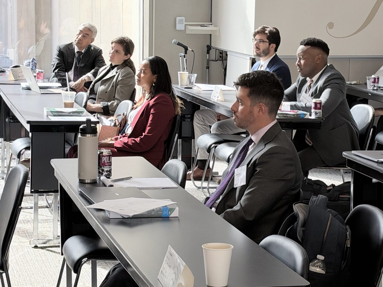 A group of professionals in business attire sitting at rows of tables in a conference room, listening to a presentation.